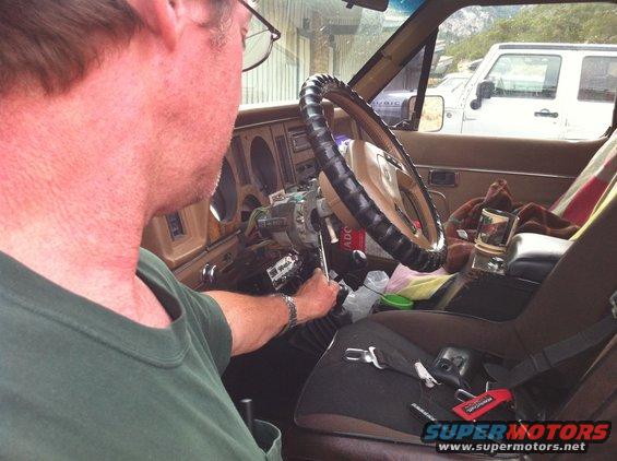 Interior of a brown vehicle with a person working on the steering column, a white Jeep visible outside