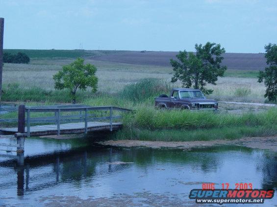 Maroon 1980s pickup truck parked next to a wooden bridge over water. Rural landscape in background