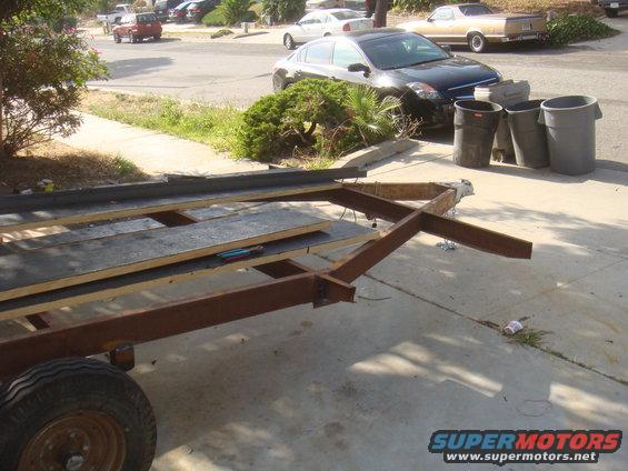 Trailer frame with wood planks, parked on driveway. Cars and trash cans in background