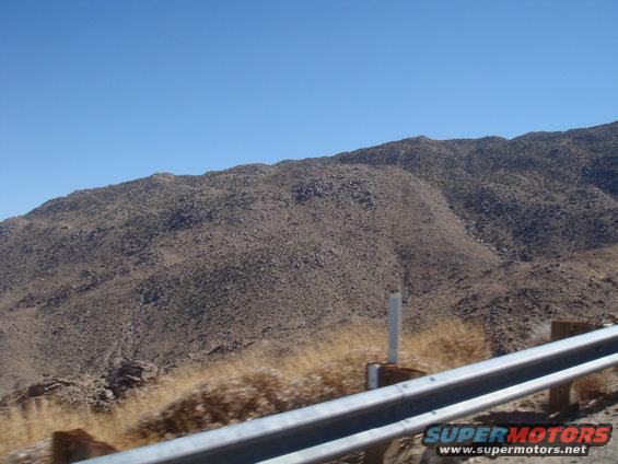 Desert mountains under a clear blue sky, with a metal guardrail in the foreground. Text 'SUPER MOTORS' is visible
