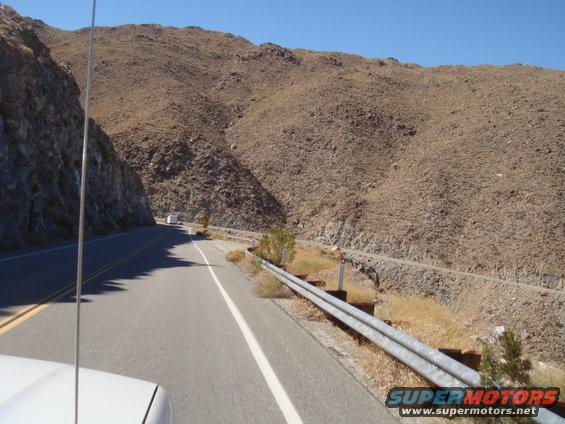 Driving on a two-lane road through a rocky, mountainous desert landscape under a clear blue sky. A white truck is visible far ahead