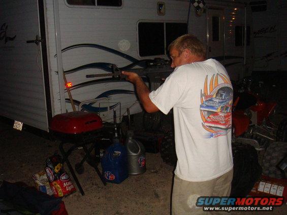 Man with blonde hair, wearing a white t-shirt with a racing graphic, holds a tool near a white trailer. A red ATV is partially visible
