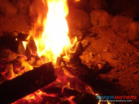 Campfire with logs and rocks, bright flames, text "SUPERMOTORS www.supermotors.net" at bottom right