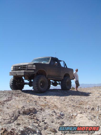 Dark 1992 Ford Bronco with large tires and roll cage, parked on a dirt hill under a clear blue sky