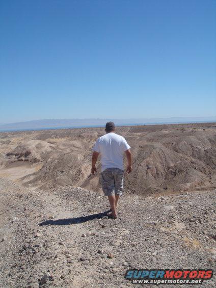 Man walks away from camera into a barren desert with mountains in the distance under a clear blue sky