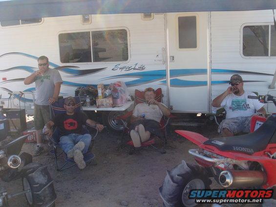Four guys chilling outside a trailer with ATVs parked nearby