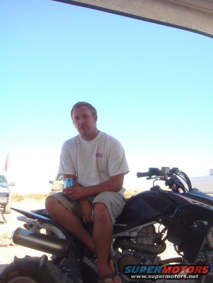 Man in t-shirt and shorts sits on black ATV, holding a can. Two trucks visible in background