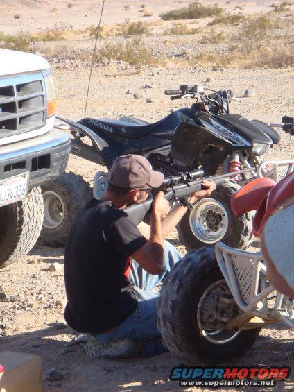 Person kneels aiming a rifle next to a black Honda ATV and part of a white Ford truck. Another ATV is visible