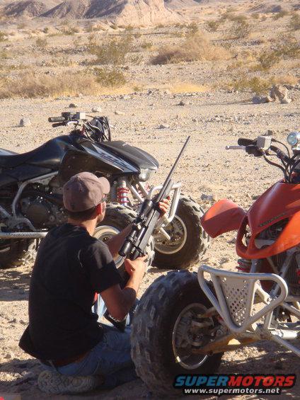 Person kneels with rifle between two ATVs in a desert landscape