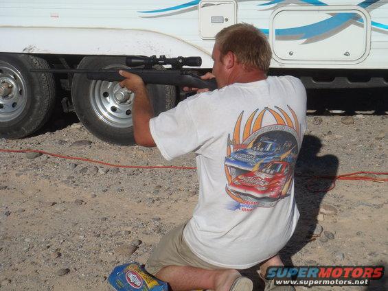 Man in white t-shirt aims rifle at the side of a white trailer with blue graphics