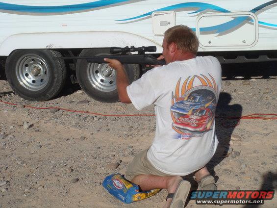Man aims rifle at side of white trailer with blue stripe