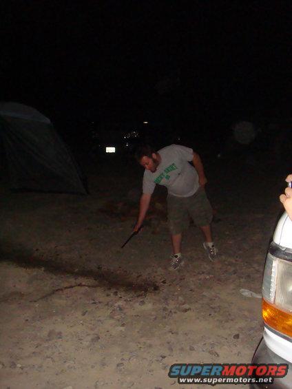 Person poking ground near a tent and the front corner of a white vehicle