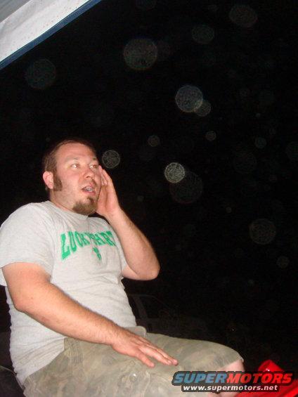 Man in a grey t-shirt and camouflage shorts sits in a vehicle at night, with blurry lights in the background