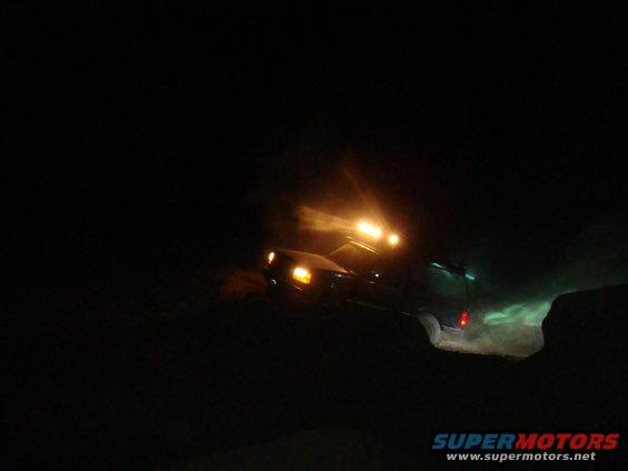 SUV with lights on driving on a dirt road at night, kicking up dust