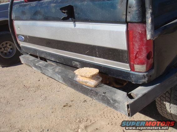 Dark green truck tailgate with silver and black stripe, cracked red taillight, and heavy-duty bumper holding a sandwich