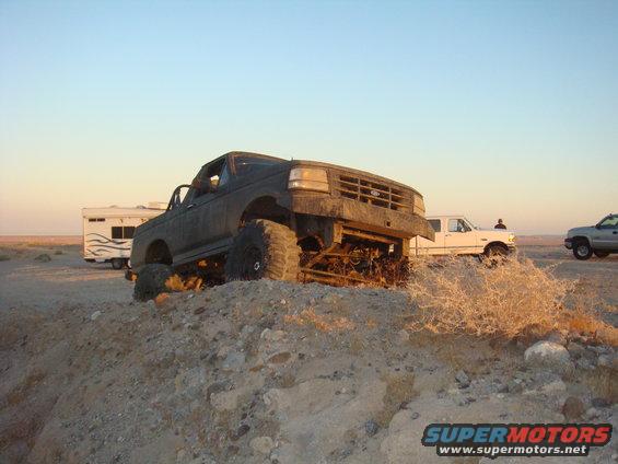 Dark SUV with big tires on a dirt mound, white trailer and truck behind it