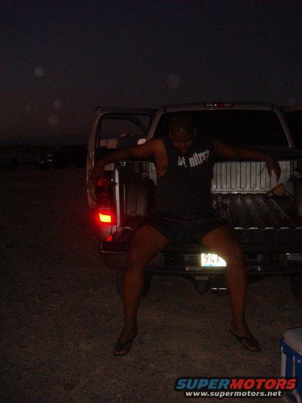 Man in shorts and t-shirt sits on the tailgate of a pickup truck at night. The truck's taillight is illuminated