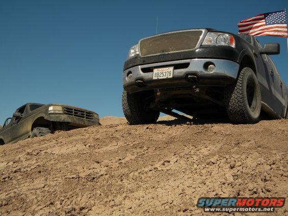 Black Ford F-150 truck with American flag, license plate 8B25378, and a dirt-covered Ford Bronco on a sandy hill
