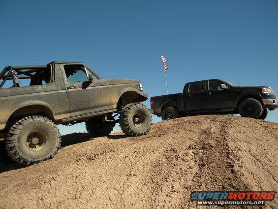 Modified SUV with large tires on a dirt hill, next to a black pickup truck