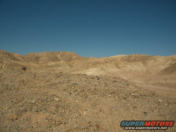 Desert hills and rocky ground under a clear blue sky. Supermotors logo in the corner