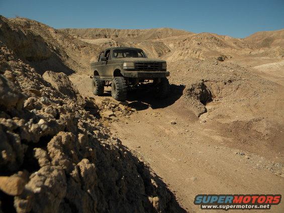 Dark 1992 Ford Bronco with lifted suspension and large tires driving on a dirt trail in a desert landscape