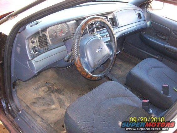 Interior shot of a Ford Crown Victoria, showing the dashboard, steering wheel, and front seats. The floor is dirty