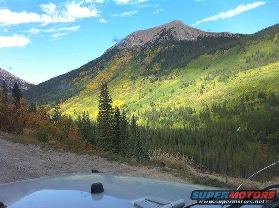 Mountain valley with green and yellow trees under a blue sky with clouds. Part of a vehicle hood is visible in the foreground