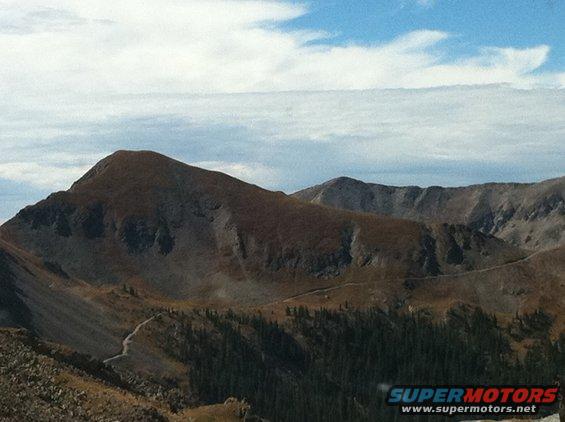 Mountain landscape with a winding road through trees under a cloudy sky. Watermark reads SUPERMOTORS.NET
