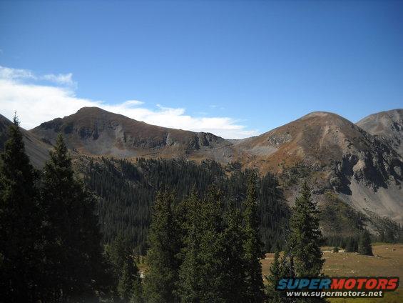 Mountain landscape with evergreen trees and a clear blue sky, a watermark reads SUPERMOTORS