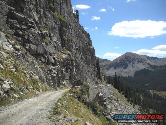 Gravel road winding along a steep mountain cliff face with trees and a distant mountain peak under a blue sky with clouds