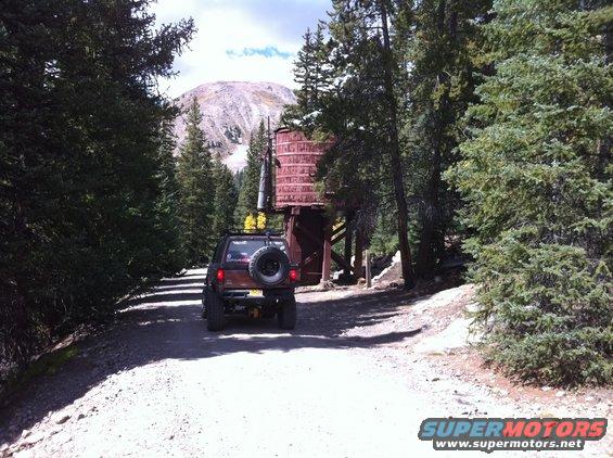 Brown SUV with a spare tire on the back driving on a dirt road near a water tower and mountain