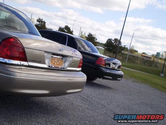 Two Ford Crown Victorias parked on gravel, rear view. One is tan, the other black. Tan car has license plate FCX-2527