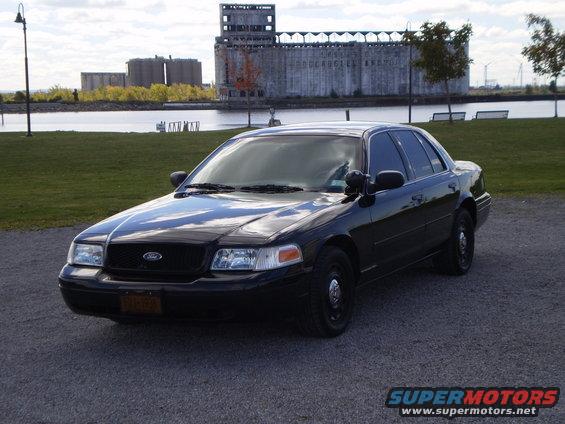 Black 2003 Ford Crown Victoria sedan parked on gravel, with industrial buildings and water in the background
