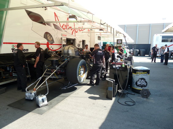 Drag racing car chassis with large rear tires and engine components, being worked on by crew members under a canopy