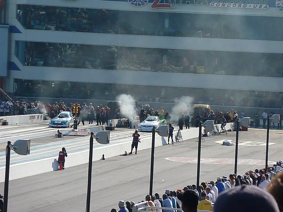 Two drag cars on a track, one blue and white, the other green and white, with smoke coming from their tires. Spectators watch from the stands