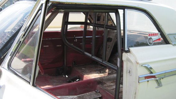 Interior of a white 1964 Ford Fairlane with a roll cage, red seats, and a Moroso sticker on the rear window