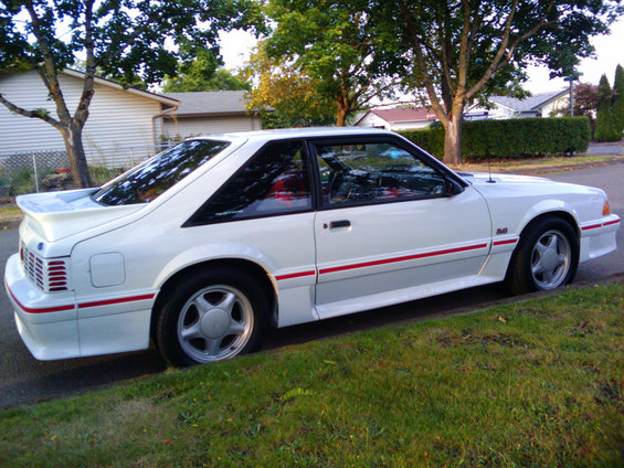 White 1987 Ford Mustang coupe parked on street, side view showing red stripe and spoiler