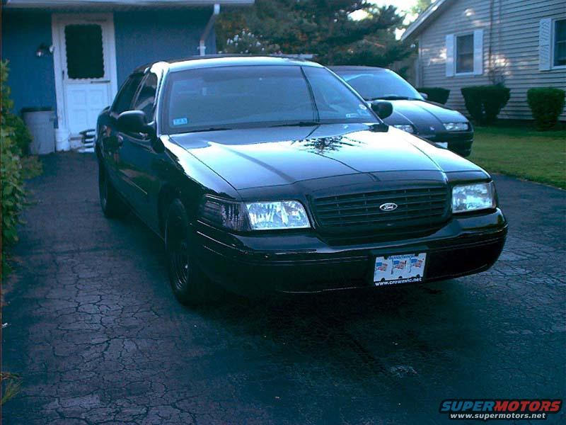 Black 2000 Ford Crown Victoria parked in a driveway, front 3/4 view, with a second dark car behind it