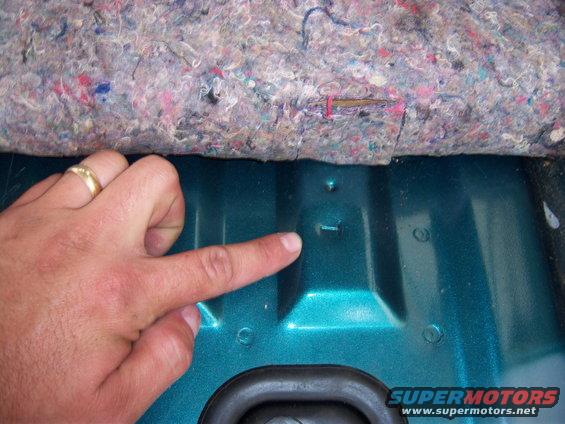 Hand pointing to a small metal tab on teal interior paneling, carpet above