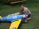 1997 Ford Deluxe - A man kneels next to a blue and yellow model airplane on a grassy lawn.