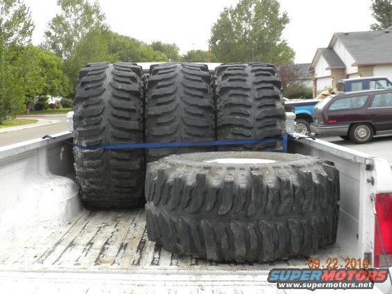 Four large off-road tires secured in a pickup truck bed. Other vehicles and houses are visible in the background