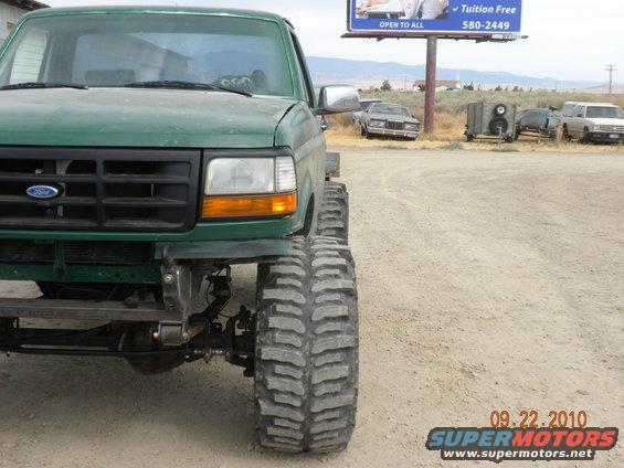 Green Ford F-Series truck with large tires, front view. Other vehicles and a billboard are in the background