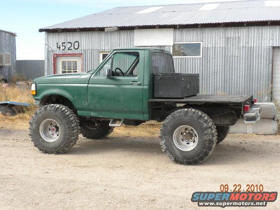 Green flatbed truck with oversized tires and a toolbox on the bed, parked in front of a corrugated metal building
