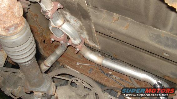 Underside of a vehicle showing exhaust pipes and a driveshaft, with rust on the frame