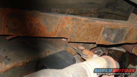 rusted frame rail with bolts, driveshaft U-joint, and exhaust pipe section visible