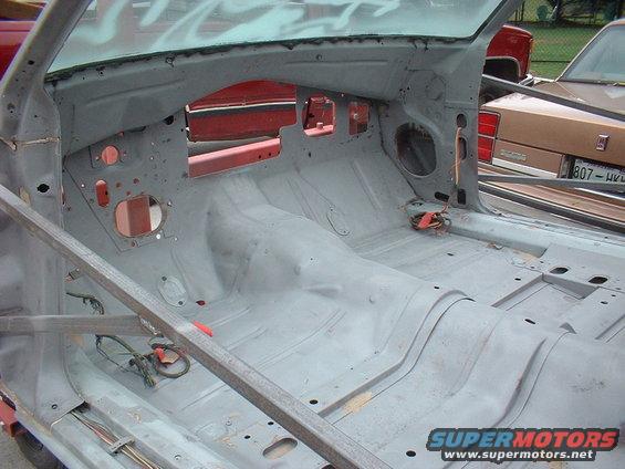 Interior of a car stripped down for restoration, showing bare metal floor and body panels. Another car is visible in the background