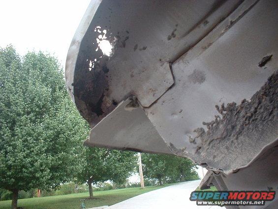 Close-up of rusted metal on a car's undercarriage, with trees in the background