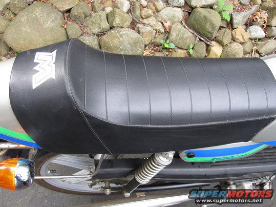 Black motorcycle seat with white 'M' logo, silver bodywork, and blue/green stripe