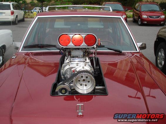 Red car with a large supercharger protruding from the hood. Other vehicles are visible in the background
