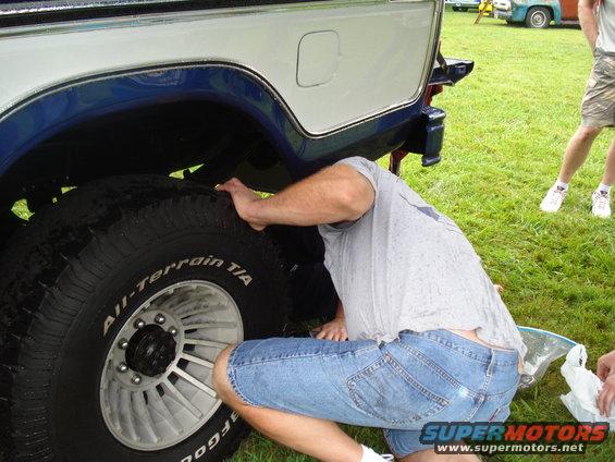 Guy checking out the rear tire of a blue and white pickup truck. Another truck is visible in the background